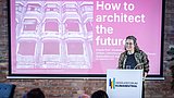 Foto, Person steht am Rednerpult mit Schriftzug 'Gebäudeforum Klimaneutral' vor Präsentationsfolie 'How to architect the future' mit Bild eines Gebäudes