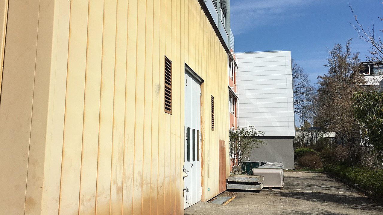Foto, Ansicht einer Gebäudeseite mit gelber Vertäfelung, einer weißen Tür und angrenzenden modernen Strukturen unter einem klaren blauen Himmel.