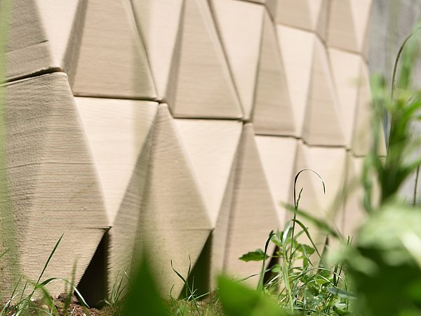 Foto, Nahaufnahme einer Fassade mit dreieckigen, reliefartigen Mustern und grünem Gras im Vordergrund.