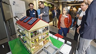 Foto, Ein detailliertes Modellhaus mit Sonnenkollektoren, umgeben von Teilnehmern, die auf einer Veranstaltung diskutieren.