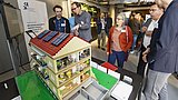Foto, Ein detailliertes Modellhaus mit Sonnenkollektoren, umgeben von Teilnehmern, die auf einer Veranstaltung diskutieren.