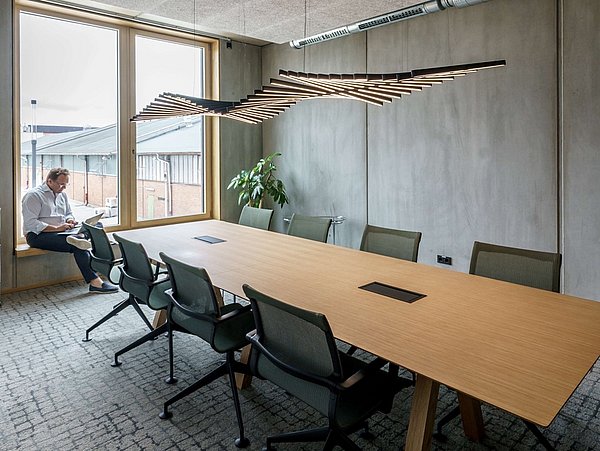 Foto, Ein Konferenzraum mit einem langen, rechteckigen Holztisch und mehreren drehbaren Bürostühlen. An der Decke hängt eine markante, wellenförmige Lampe aus Holzleisten. Große Fenster lassen Tageslicht herein und bieten einen Blick auf ein Industriegebäude. Eine Person sitzt am Fenster und arbeitet mit einem Laptop. Die Wände sind grau und wirken betonartig, der Boden ist mit einem hellen Teppich ausgelegt. Ein weißes Sideboard steht an der Wand neben dem Fenster, eine grüne Pflanze befindet sich in einer Ecke.