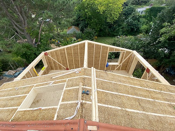 Foto, Dachkonstruktion eines Neubaus aus Stroh und Holz im Modulbau, umgeben von Bäumen und Garten