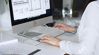 Foto, Person in weißem Hemd tippt auf einer Tastatur vor einem Bildschirm mit technischen Zeichnungen.