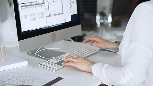 Foto, Person in weißem Hemd tippt auf einer Tastatur vor einem Bildschirm mit technischen Zeichnungen.