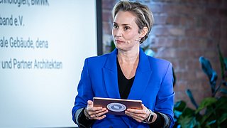 Foto, Frau in blauem Blazer hält Tablet und steht vor Präsentationsbildschirm mit Text in einem Veranstaltungsraum