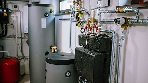 Foto, technischer Heizungsraum mit einem grauen Warmwasserspeicher, mehreren Pumpen und einem roten Ausdehnungsgefäß. Im Hintergrund sind komplexe Rohrleitungen, Ventile und Messgeräte zu erkennen, die an der Wand montiert sind..