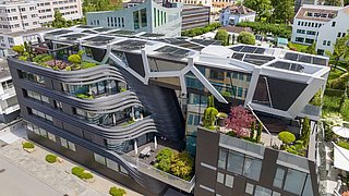 Foto, Luftaufnahme eines modernen, einzigartig gestalteten Gebäudes mit Sonnenkollektoren und grünen Dachgärten, umgeben von üppiger Vegetation.