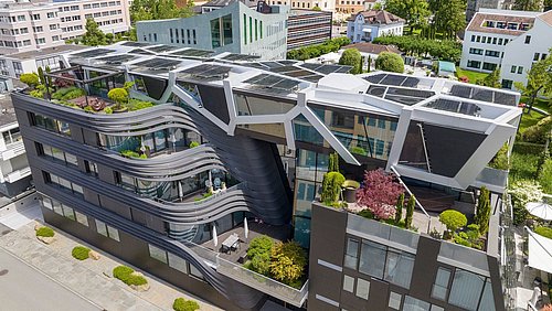 Foto, Luftaufnahme eines modernen, einzigartig gestalteten Gebäudes mit Sonnenkollektoren und grünen Dachgärten, umgeben von üppiger Vegetation.
