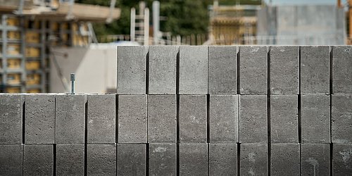 Foto, graue Betonsteine mit einer Schraube in der oberen Reihe, im Hintergrund eine Baustelle mit Gerüsten und Bäumen.