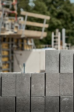 Foto, graue Betonsteine mit einer Schraube in der oberen Reihe, im Hintergrund eine Baustelle mit Gerüsten und Bäumen.