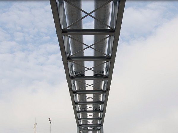 Unterseite einer Stahlbrücke, die Rohrleitungen trägt, mit Gitterträgern vor bewölktem Himmel.