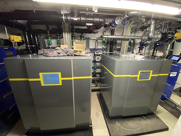 Foto, Zwei große graue Inneneinheiten von Wärmepumpen mit gelben Linien und Displays in einem Technikraum mit Rohren und Ventilen