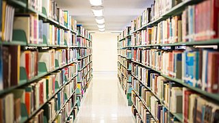 Foto, Blick in einen Gang mit Bücherregalen auf der linken und rechten Seite. 