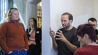 Foto, Gruppe von Menschen in einem Raum, eine Person gestikuliert mit den Händen, andere hören zu