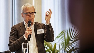 Foto, ein Redner gestikuliert leidenschaftlich, während er ein Mikrofon in der Hand hält und an einem Podium mit Wassergläsern und Pflanzen im Hintergrund steht.