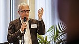 Foto, ein Redner gestikuliert leidenschaftlich, während er ein Mikrofon in der Hand hält und an einem Podium mit Wassergläsern und Pflanzen im Hintergrund steht.