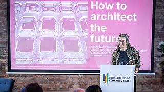 Foto, Person steht am Rednerpult mit Schriftzug 'Gebäudeforum Klimaneutral' vor Präsentationsfolie 'How to architect the future' mit Bild eines Gebäudes