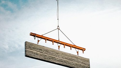 Foto, Nahaufnahme eines Betonelements, welches an einem Kran vor blauem Himmel hängt.