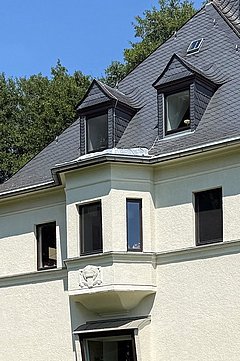 Foto, Ein helles, zweistöckiges Wohnhaus mit dunklem Schieferdach steht auf einer gepflegten Rasenfläche. Die Fassade ist cremefarben, das Sockelgeschoss aus rötlichem Stein. Mehrere Fenster mit dunklen Rahmen sind sichtbar, einige davon im Dachbereich mit Gauben.