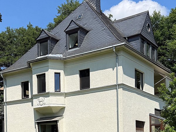 Foto, Ein helles, zweistöckiges Wohnhaus mit dunklem Schieferdach steht auf einer gepflegten Rasenfläche. Die Fassade ist cremefarben, das Sockelgeschoss aus rötlichem Stein. Mehrere Fenster mit dunklen Rahmen sind sichtbar, einige davon im Dachbereich mit Gauben.