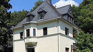 Foto, Ein helles, zweistöckiges Wohnhaus mit dunklem Schieferdach steht auf einer gepflegten Rasenfläche. Die Fassade ist cremefarben, das Sockelgeschoss aus rötlichem Stein. Mehrere Fenster mit dunklen Rahmen sind sichtbar, einige davon im Dachbereich mit Gauben.