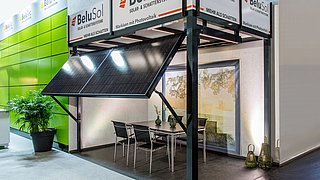 Foto, ein moderner Messestand mit Sonnenkollektoren, einem Esstisch mit Stühlen und dekorativen Pflanzen, der nachhaltiges Design präsentiert.