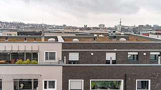 Foto, Dachflächen und Fassaden moderner Wohngebäude mit Fenstern und Rollläden, im Hintergrund Stadt mit Kirchturm und weiteren Gebäuden.