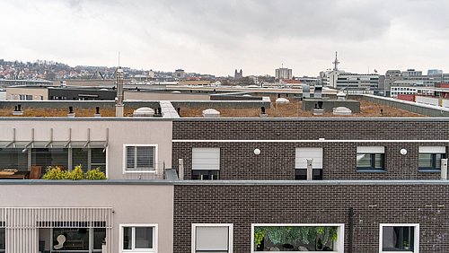 Foto, Dachflächen und Fassaden moderner Wohngebäude mit Fenstern und Rollläden, im Hintergrund Stadt mit Kirchturm und weiteren Gebäuden.