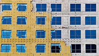 Foto, frontaler Blick auf eine mehrstöckige Neubaufassade. Die Hälfte der Fassade ist bereits mit Dämmplatten versehen, die andere Hälfte ist noch im Rohbau.