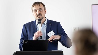 Foto, Mann in blauem Sakko hält Mikrofon und steht hinter einem schwarzen Rednerpult mit offenem Laptop.