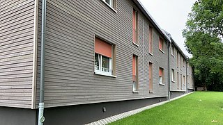 Foto, Seitenansicht eines modernen Holzgebäudes mit orangefarbenen Fensterläden, umgeben von grünem Gras.