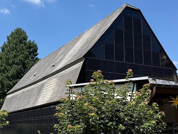 Foto, Ein großes, traditionelles Scheunengebäude mit steilem Satteldach steht im Vordergrund. Die Dach- und Fassadenflächen sind vollständig mit dunklen Solarmodulen bedeckt. Vor dem Gebäude wachsen mehrere kleine Laubbäume, deren Blätter einen Teil der Fassade verdecken.