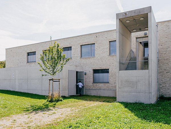 Flaches, zweigeschossiges Gebäude aus hellem Sichtbeton und Ziegeln mit kleinem Baum davor und Person an der Tür, Technikzentrale des Kalten Nahwärmenetzes.