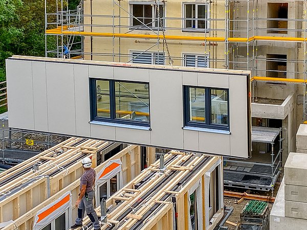 Foto, Kran hebt vorgefertigte Gebäudewand, im Hintergrund Baustelle mit Gerüst und Rohbau