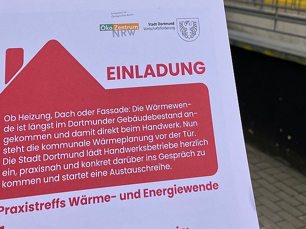 Foto, Hand hält Einladung mit Hausumriss und Text "Praxistreff", im Hintergrund Gehweg mit Geländer.