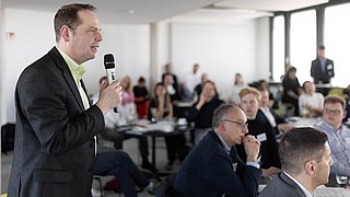 Foto, Mann im Anzug hält Mikrofon und spricht vor sitzendem Publikum in einem hellen Konferenzraum.