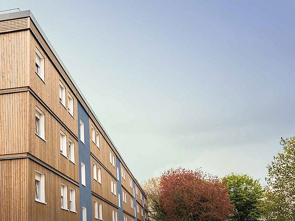 Foto, links befindet sich ein Mehrfamilienhaus mit Holzfassade. Rechts im Bild hält sich eine große Gruppe von Menschen vor dem Haus auf.