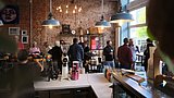Foto, Innenraum eines Cafés mit mehreren Personen, die an der Theke und im Gastraum stehen, sichtbare Kaffeemaschine und große Fenster.