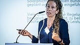 Foto, Frau mit lockigem Haar hält Präsentation an einem Rednerpult mit Mikrofon, im Hintergrund Text zur Solarpflicht in Deutschland