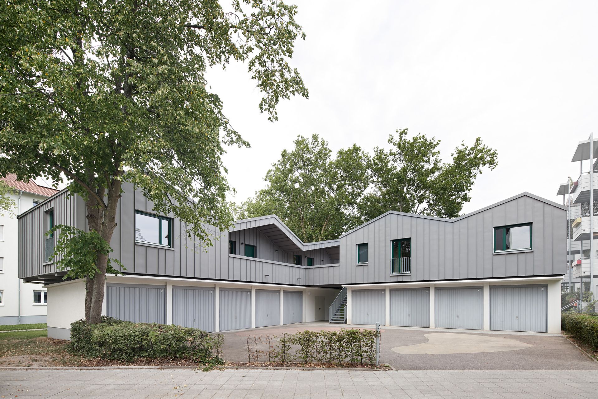 Foto, Moderne Wohngebäude mit grauen Fassaden und Garagen im Erdgeschoss, umgeben von Bäumen und Gehweg
