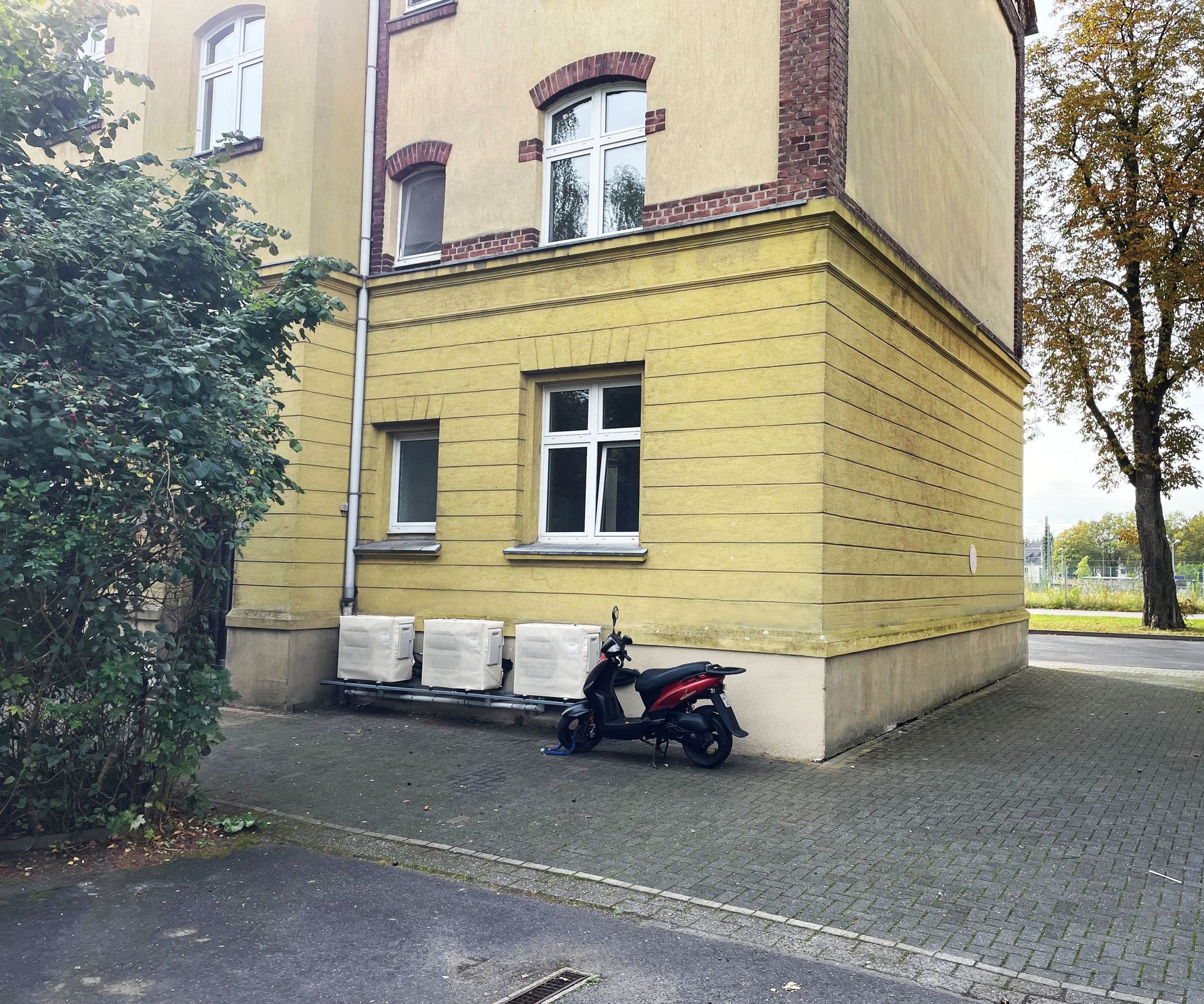 Foto, Gelbes Wohngebäude mit weißen Fenstern, davor ein schwarzer und roter Motorroller auf gepflastertem Hof