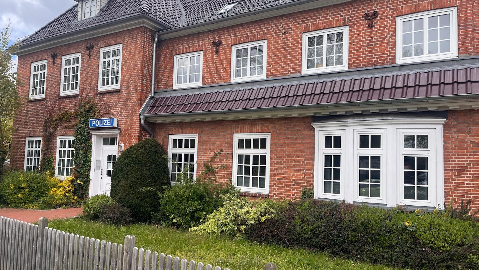 Foto, Zweistöckiges rotes Backsteingebäude mit mehreren weißen Fenstern, einem Schild mit der Aufschrift 'POLIZEI' und einem kleinen Garten mit Sträuchern davor.