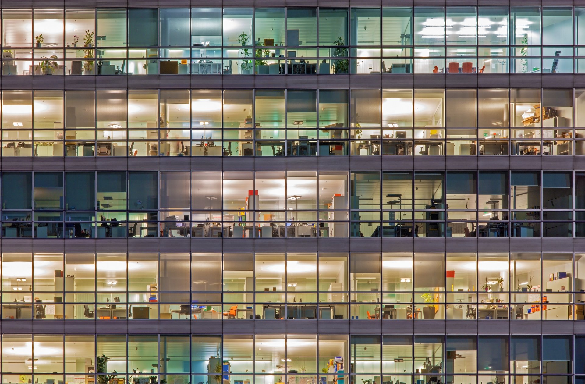Foto, frontaler Blick auf die vollständig verglaste Fassade eines Bürogebäudes bei Dunkelheit. In den beleuchteten Räumen sind Einrichtungsgegenstände zu erkennen.