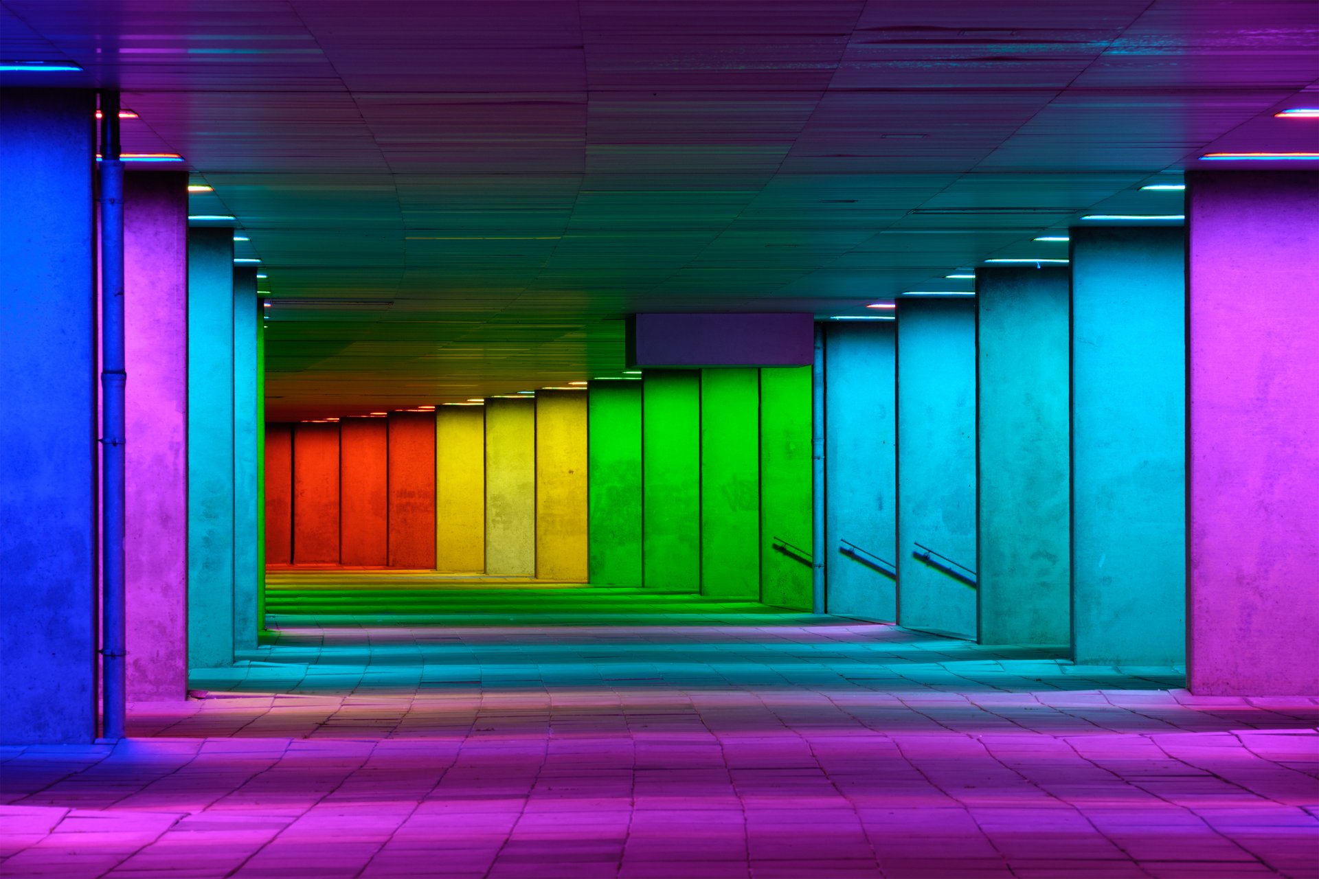 Foto, langer Gang mit Betonwänden, rechts gehen mehrere Treppenaufgänge ab, die in verschiedenen Farben beleuchtet sind.