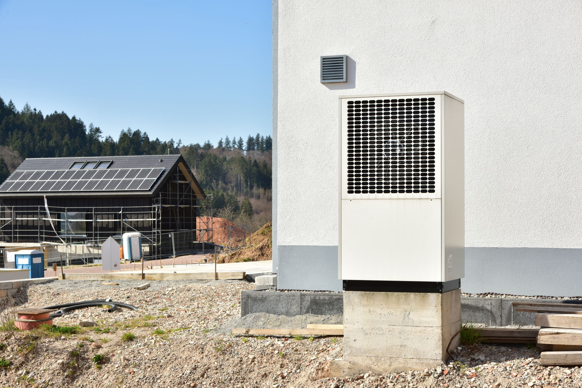 Foto, im Vordergrund ein Haus mit davorstehender Wärmepumpe, im Hintergrund ein Haus im Bau mit Solarkollektoren auf dem Dach.