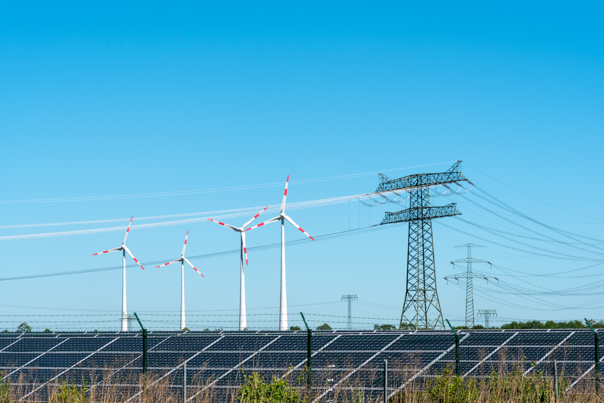 Foto, im Vordergrund eine Solaranlage auf einer Wiese, im Hintergrund Windräder und ein Strommast.