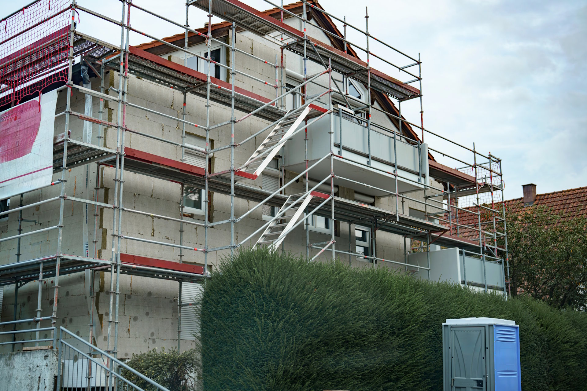 Foto, Mehrstöckiges Wohnhaus mit Gerüst und Baustellenaufzug, davor dichter grüner Busch und mobiler Toilettencontainer