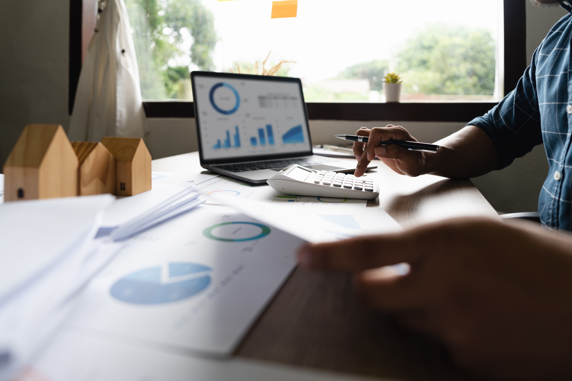 Foto, ein Mann sitzt an einem Schreibtisch und tippt auf einem Taschenrechner, im Hintergrund werden auf einem Computermonitor verschiedene Diagramme dargestellt.