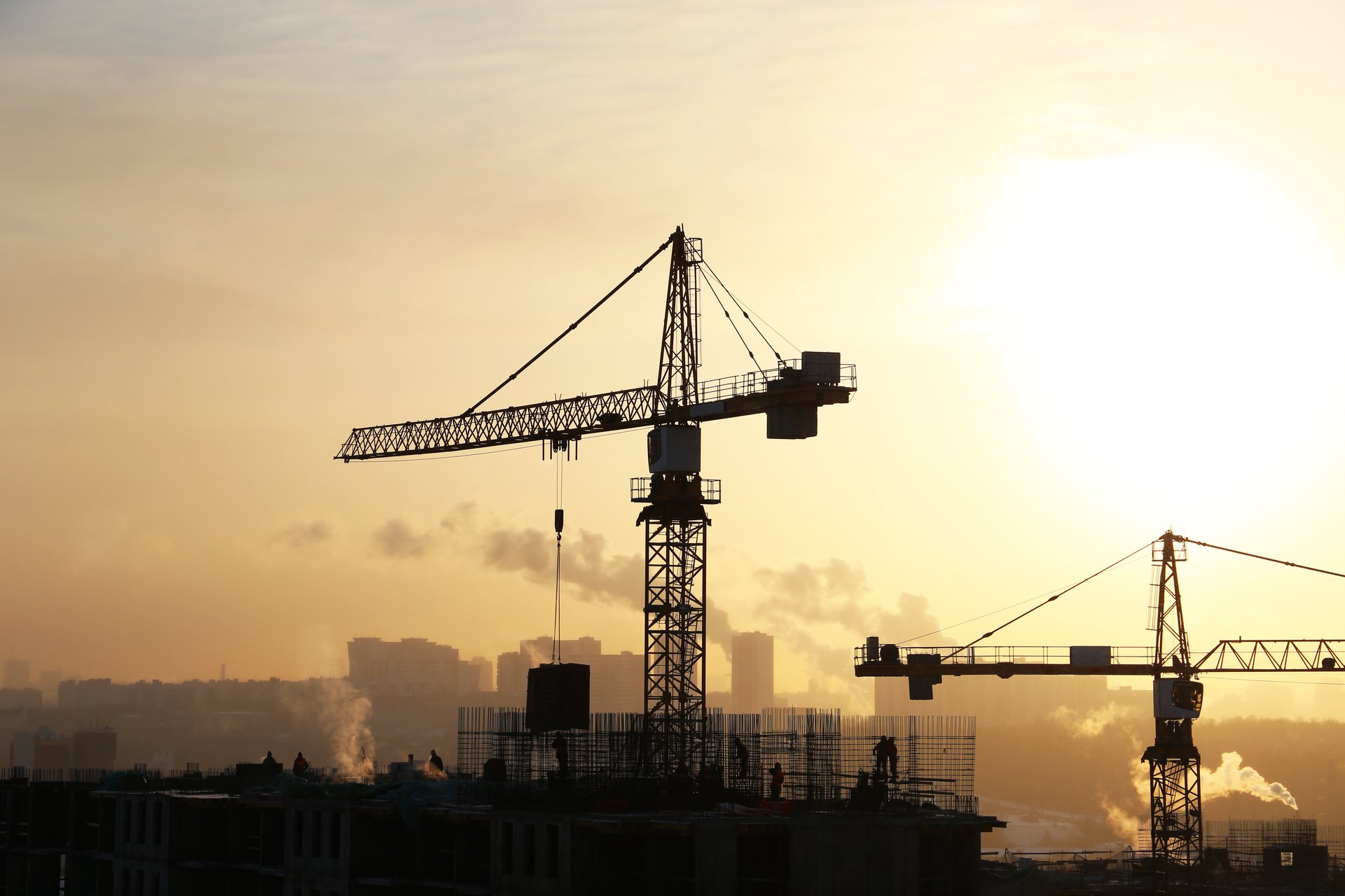 Foto, zwei Kräne sowie ein Rohbau als schwarze Silhouetten im Gegenlicht der Sonne. Am Horizont rauchende Fabrikschornsteine.